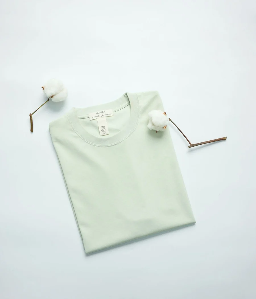 A folded pale green cotton tee on a light surface with cotton stems